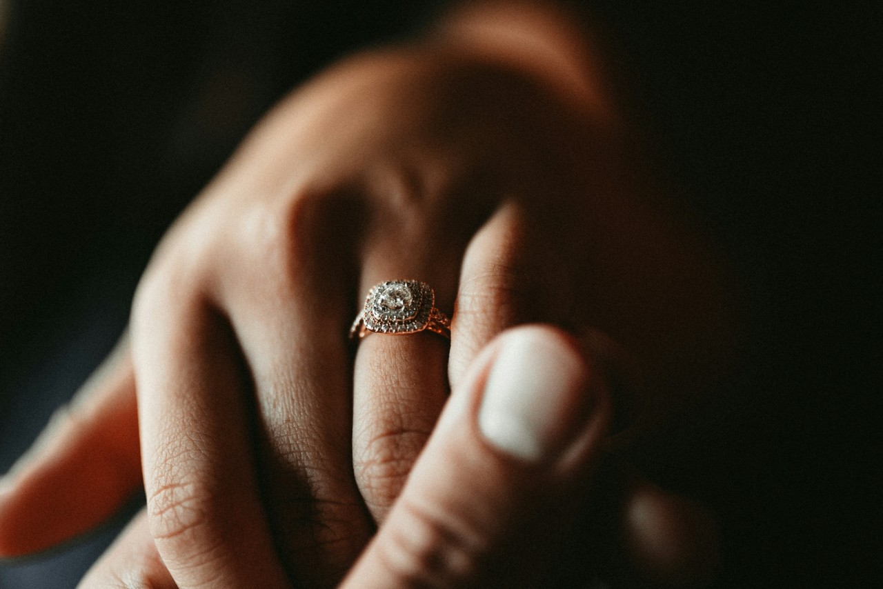 a man’s hand holding a woman’s, which is adorned with a double halo engagement ring