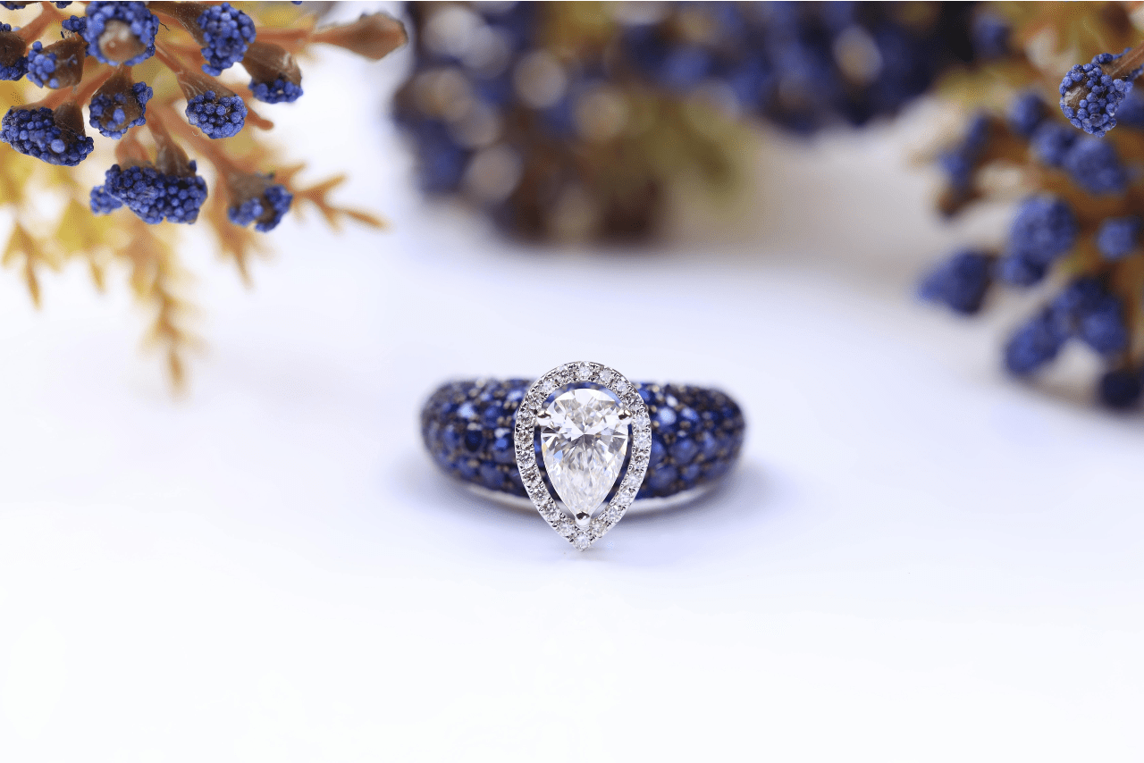 A unique engagement ring with a pear shape halo setting and blue side stones displayed among dried juniper branches.